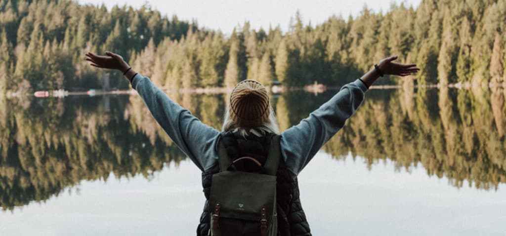 Happy woman with arms raised in air standing in front of lake surrounded by trees