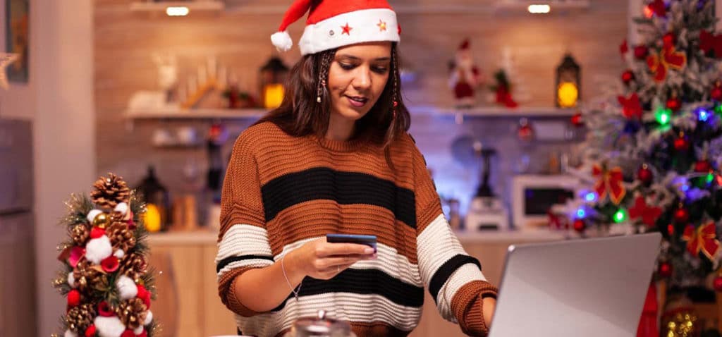 Woman wearing Santa hat surrounded by Christmas decorations, looking down at credit card in her hand while paying online