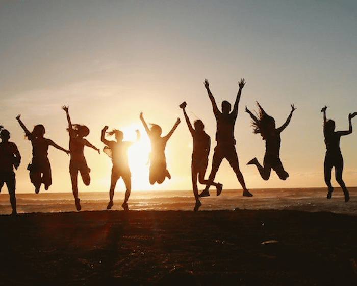 Group of people jumping in air with hands and feet outright on top of mountain in front of sunset